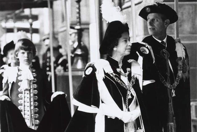 The Silver Jubilee visit in May 1977 also saw the Queen and the Duke of Edinburgh attend an Order of the Thistle service in St Giles' Cathedral. 
Here, they are walking along a covered walkway to the cathedral. They both wear ceremonial robes and chains around their necks, and black caps with large white feather plumes. The train from the Queen's gown is held up by a page boy who is walking behind her.