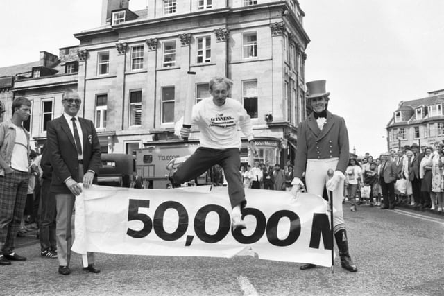 British athlete David Hemery leaps the 50,000 mile hurdle with the Friendship Scroll, which had journeyed through 25 countries ahead of the Commonwealth Games 1986. Holding the banner (left) is former Lord Provost Kenneth Borthwick, Chairman of the Commonwealth Games executive.