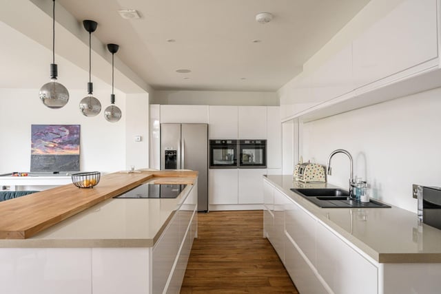 The contemporary fully-fitted kitchen area with a range of integrated white goods, breakfast bar and under-unit lighting,  finished with stylish white units and a wooden worktop.
