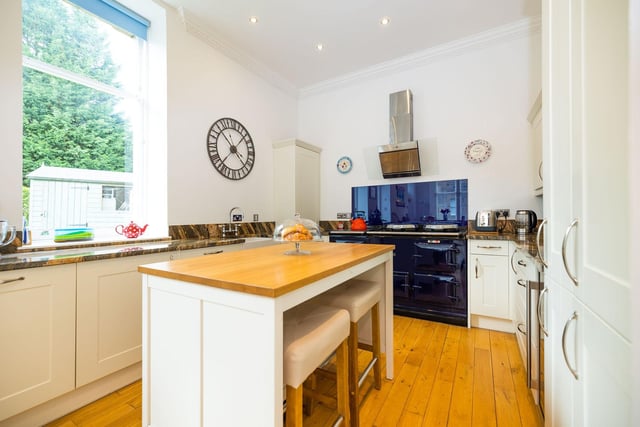 The spacious dining kitchen, boasting floor-to-ceiling windows, wooden flooring, and pendant lighting. White shaker-style wall and floor units sit alongside marble worktops, a Belfast sink, and integrated appliances including an AGA, a fridge/freezer, integrated dishwasher, washing machine and wine cooler.
