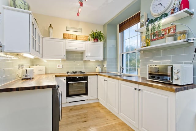 The Loanhead property's kitchen features a trio of southwest-facing windows (one with a charming window seat) which capture sunny natural light throughout the day and frame leafy views of the adjacent green space. Attractive white wall and base cabinets are accompanied by rich wood-styled worktops and chic metro-tiled splashbacks. An oven and hob are integrated, whilst provision is made for additional freestanding and undercounter appliances.