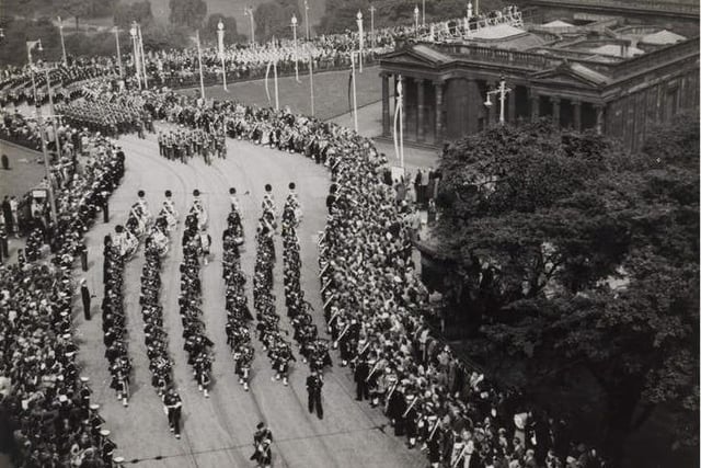 Another picture of the procession with the Honours of Scotland on June 24, 1953, shows the military parade marching up the Mound past the National Gallery.  
Crowds have gathered on both sides of the road to watch the spectacle. Military personnel in naval uniform stand at the edge on either side of the road and separate the spectators from the procession. Tall poles with banners attached have been placed at regular intervals in the grounds of the gallery along the route of the procession.