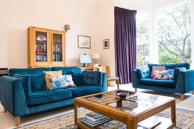 The property's living room, with bay windows overlooking the private garden. Boasting high ceilings, cast iron radiators, and a soft colour palette including carpeting, and a focal mantle housing a log-effect gas stove.