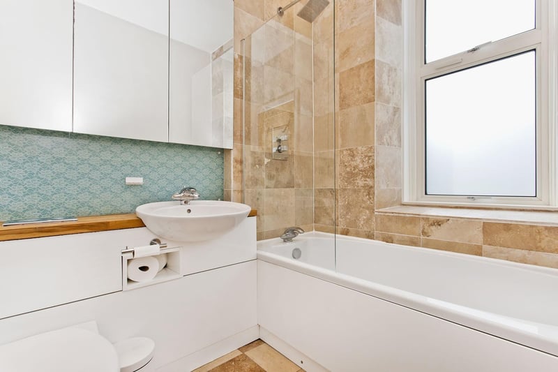 The contemporary bathroom, with stylish tiling and natural travertine flooring, comprises a bath with a rainfall showerhead, a WC-suite set into a vanity unit, and underfloor heating.