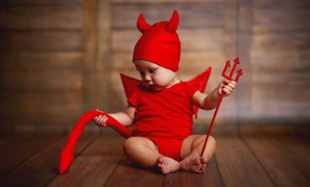 A baby in a devil halloween costume with horns and trident on a dark wooden background